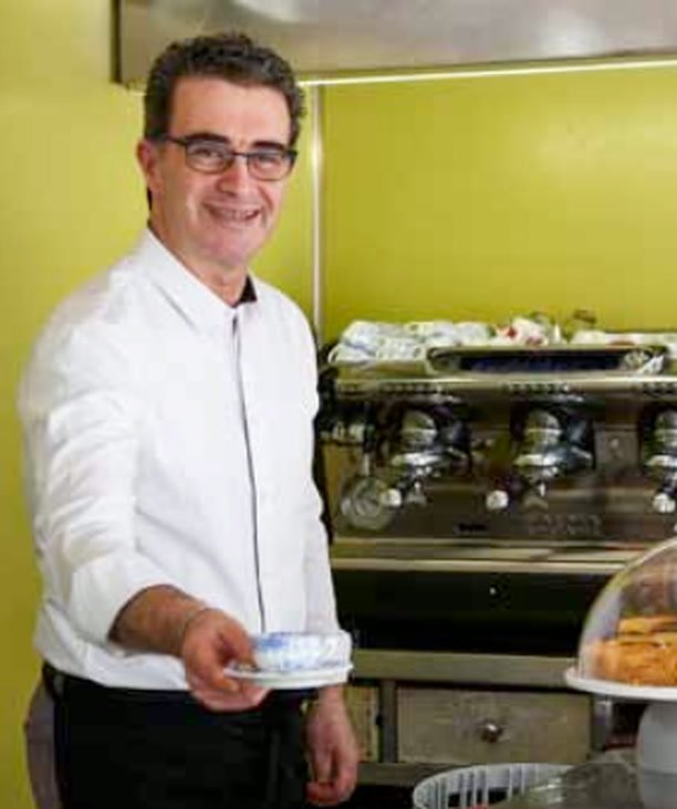 Dietro il bancone, in camicia bianca, con una tazzina in mano davanti alla macchina del caffè